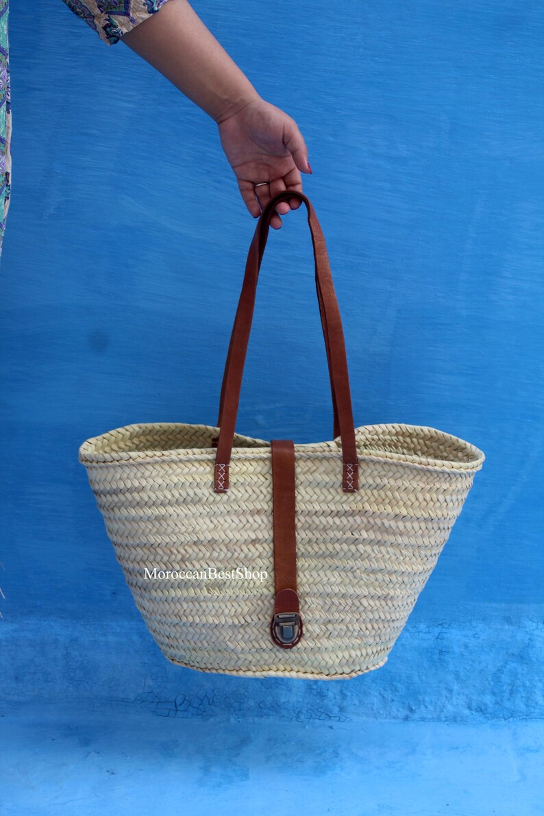 May include: A woven straw tote bag with brown leather handles and accents. The bag has a wide opening and a rectangular base. The handles are attached with decorative stitching. The bag is held against a blue background. The text "MoroccanBestShop" is visible on the bag.