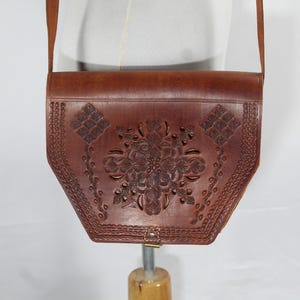 Puede incluir: Bolso bandolera de cuero marrón con diseño floral. El bolso tiene cierre de solapa y correa larga. La parte delantera está decorada con un estampado floral en relieve y bordes decorativos. El bolso se exhibe en un maniquí.