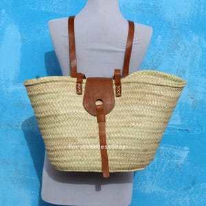 May include: A woven straw basket bag featuring two brown leather shoulder straps and a central brown leather flap closure with a vertical strap. The bag has a light tan natural fiber texture and includes the text MoroccanBestShop on the front.