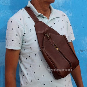 May include: A brown leather sling bag featuring a single adjustable shoulder strap, a vertical zippered front pocket with a metallic zipper pull, and visible white contrast stitching along the edges and seams of the bag's main body.