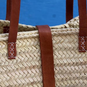 May include: Close-up of a woven straw tote bag with brown leather handles and accents. The handles are attached with white cross-stitch details. The bag is set against a blue background.