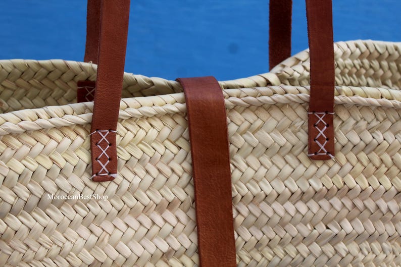 May include: Close-up of a woven straw tote bag with brown leather handles and accents. The handles are attached with white cross-stitch details. The bag is set against a blue background.