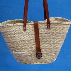 May include: Woven straw tote bag with brown leather handles and a securing strap. The bag has a natural beige color and a trapezoidal shape. The leather strap features a metal clasp and is stitched to the bag. The bag is designed for carrying items.