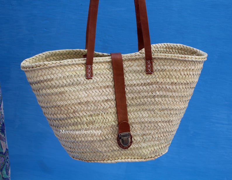 May include: Woven straw tote bag with brown leather handles and a securing strap. The bag has a natural beige color and a trapezoidal shape. The leather strap features a metal clasp and is stitched to the bag. The bag is designed for carrying items.