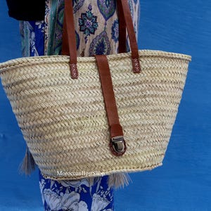 May include: Woven straw tote bag with brown leather handles and a matching strap closure. The bag has a rectangular shape and is held against a blue background. The bag is a natural color and has a metal clasp.