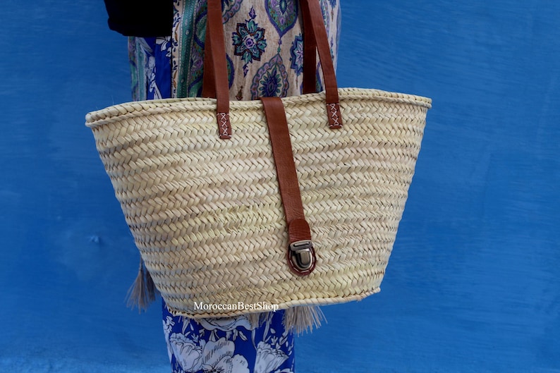 May include: Woven straw tote bag with brown leather handles and a matching strap closure. The bag has a rectangular shape and is held against a blue background. The bag is a natural color and has a metal clasp.