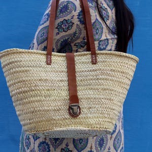 May include: A woven straw tote bag with brown leather handles and a matching strap closure. The bag is a natural beige color and has a rectangular shape. The background is a solid blue.