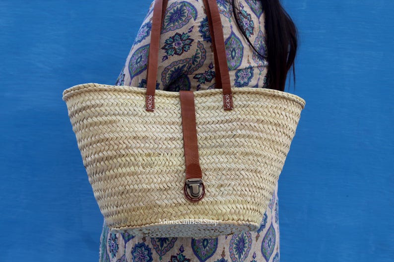 May include: A woven straw tote bag with brown leather handles and a matching strap closure. The bag is a natural beige color and has a rectangular shape. The background is a solid blue.