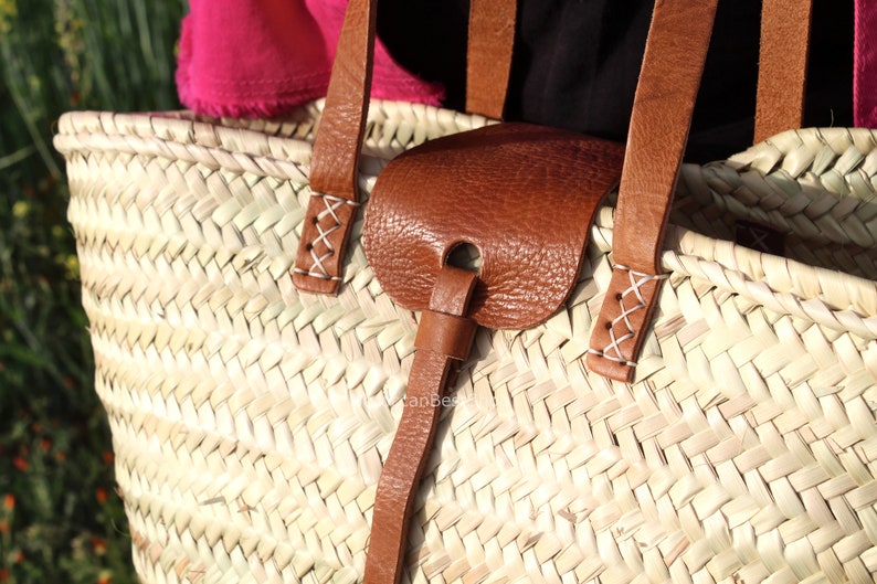 May include: A close-up of a woven straw tote bag with brown leather straps and a leather handle. The straps are stitched with white thread.