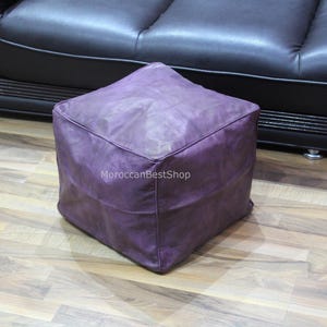 May include: A purple leather pouf ottoman with a square shape. The pouf is sitting on a wooden floor.