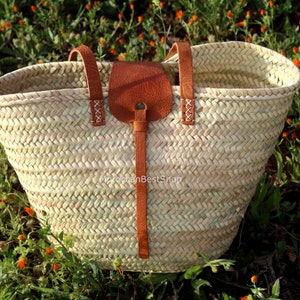 May include: A large, woven straw tote bag with brown leather handles and a leather strap. The bag is sitting on a bed of green grass with orange flowers.