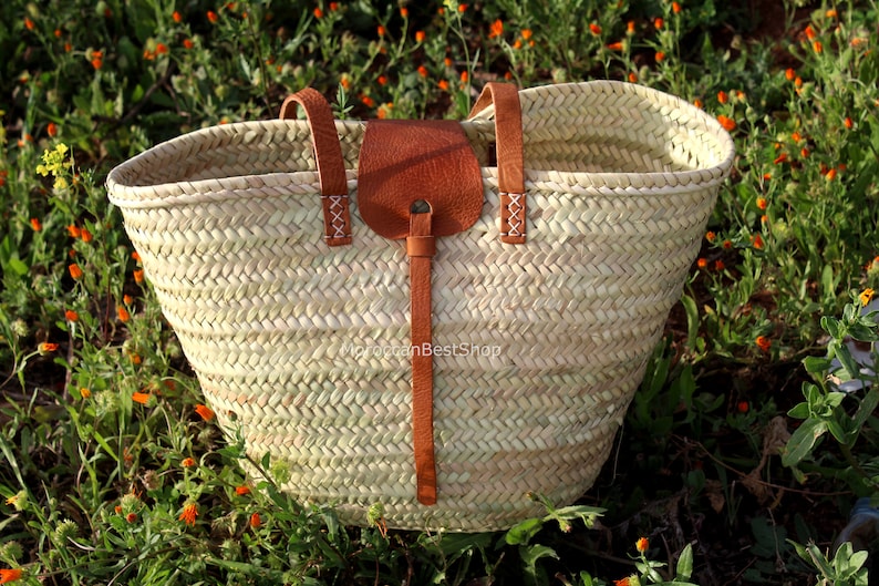 May include: A large, woven straw tote bag with brown leather handles and a leather strap. The bag is sitting on a bed of green grass with orange flowers.