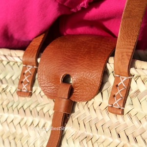 May include: Close-up of a woven straw tote bag with brown leather handles and a leather strap. The handles are stitched with white thread.