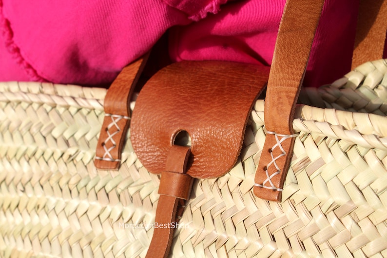 May include: Close-up of a woven straw tote bag with brown leather handles and a leather strap. The handles are stitched with white thread.