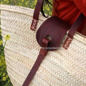 May include: A large, woven straw tote bag with brown leather straps and a brown leather handle. The bag is being held by a person wearing a red shirt.