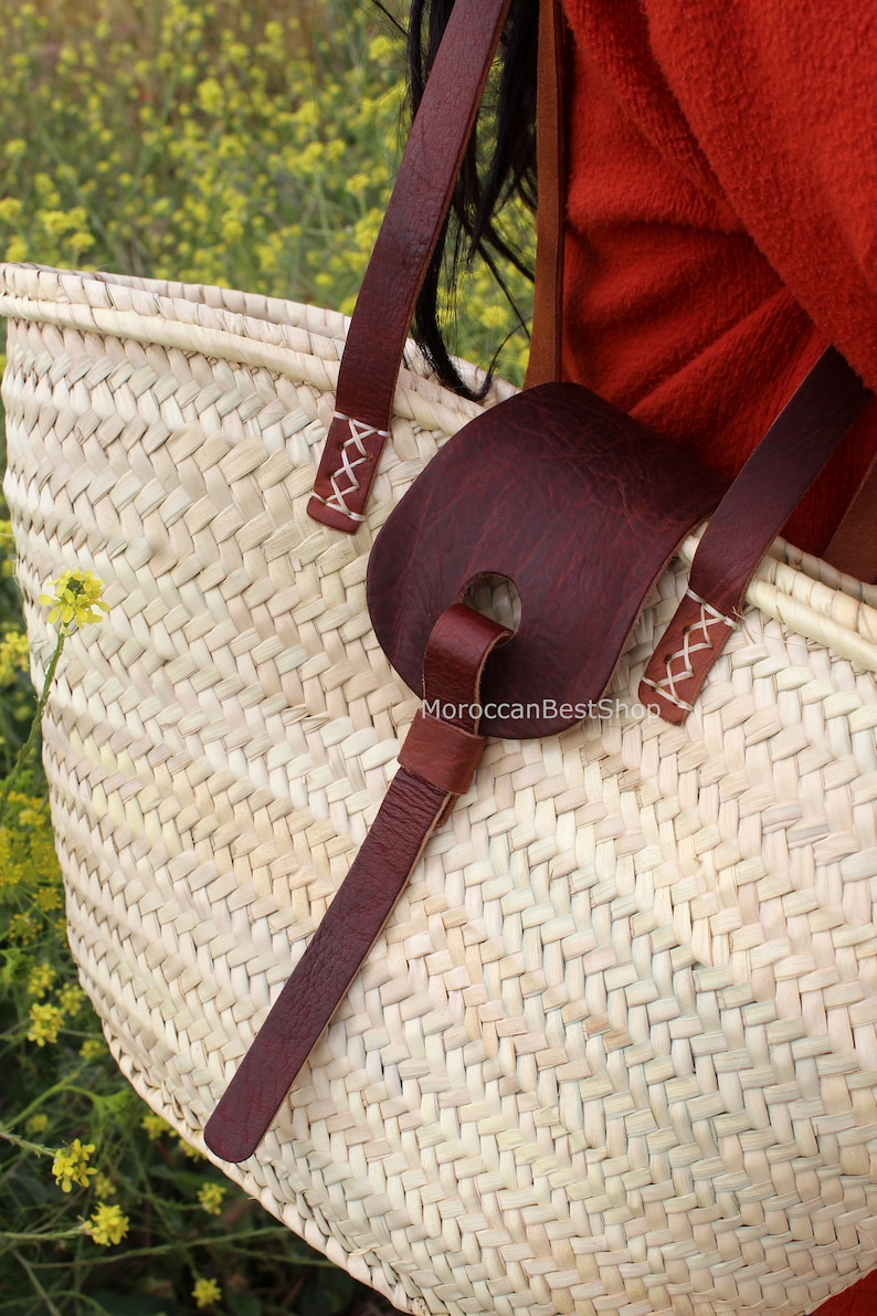 May include: A large, woven straw tote bag with brown leather straps and a brown leather handle. The bag is being held by a person wearing a red shirt.