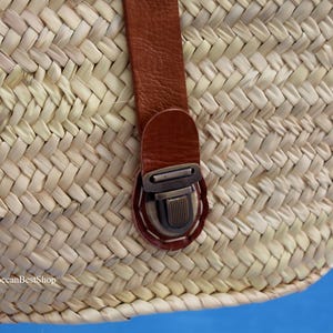 May include: Close-up of a woven straw basket with a brown leather strap and metal clasp. The basket is made of natural-colored straw and has a textured surface. The strap is attached with white stitching.
