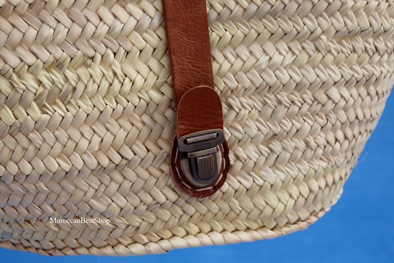 May include: Close-up of a woven straw basket with a brown leather strap and metal clasp. The basket is made of natural-colored straw and has a textured surface. The strap is attached with white stitching.