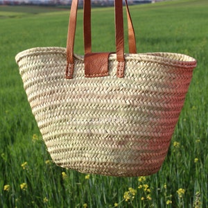 May include: A large, light brown woven tote bag with brown leather handles and a leather patch on the front. The bag is hanging in front of a green grassy field.