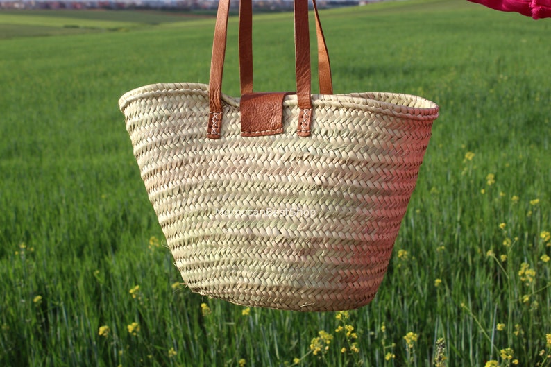 May include: A large, light brown woven tote bag with brown leather handles and a leather patch on the front. The bag is hanging in front of a green grassy field.