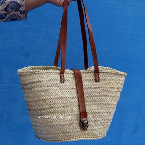 May include: A woven straw tote bag with brown leather handles and a matching strap closure. The bag has a rectangular shape with a wider top and a narrower base. The text "MoroccanBestShop" is visible at the bottom.