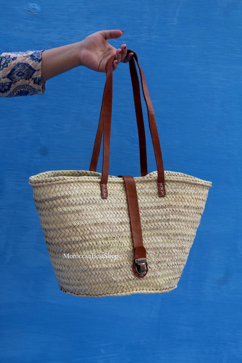 May include: A woven straw tote bag with brown leather handles and a matching strap closure. The bag has a rectangular shape with a wider top and a narrower base. The text "MoroccanBestShop" is visible at the bottom.