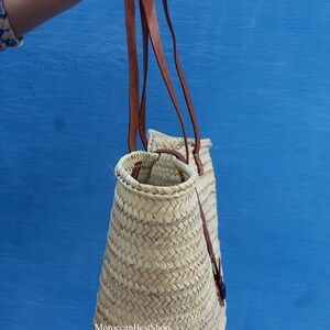 May include: A woven straw tote bag with brown leather straps. The bag has a conical shape and is held against a blue background. The text "MoroccanBestShop" is visible on the bag.