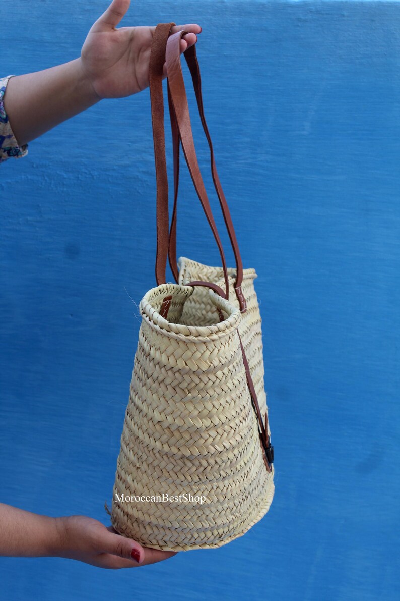 May include: A woven straw tote bag with brown leather straps. The bag has a conical shape and is held against a blue background. The text "MoroccanBestShop" is visible on the bag.