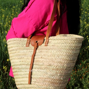 May include: A large, woven straw tote bag with brown leather straps and a brown leather handle. The bag is being held by a person wearing a pink shirt and blue jeans.