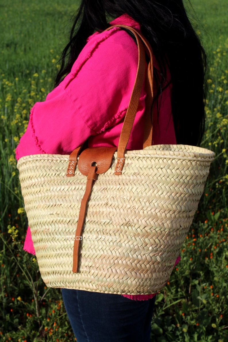 May include: A large, woven straw tote bag with brown leather straps and a brown leather handle. The bag is being held by a person wearing a pink shirt and blue jeans.