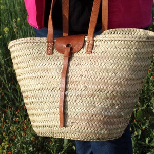 May include: A large, woven straw tote bag with brown leather handles and a leather strap with a buckle. The bag is in a field of green grass and yellow flowers.