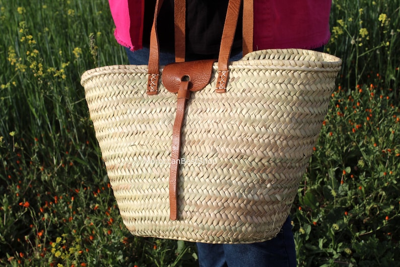 May include: A large, woven straw tote bag with brown leather handles and a leather strap with a buckle. The bag is in a field of green grass and yellow flowers.