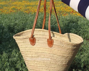 May include: A woven straw tote bag with brown leather handles and accents. The bag is held up against a backdrop of a field of yellow flowers and green foliage. The bag has a wide opening and a sturdy design.