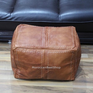 May include: A square, brown leather ottoman with white stitching. The ottoman is made of four large, stitched panels. The background includes a black leather sofa and a wooden floor. The text "MoroccanBestShop" is visible on the front of the ottoman.