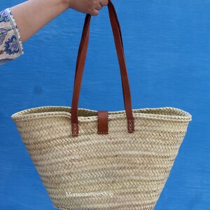 May include: A woven straw tote bag with brown leather handles and a matching leather closure. The bag is held up against a bright blue background. The bag has a rectangular shape with a wide opening.