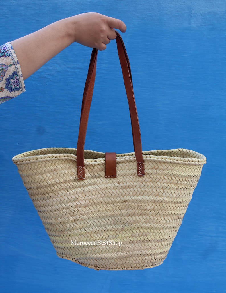 May include: A woven straw tote bag with brown leather handles and a matching leather closure. The bag is held up against a bright blue background. The bag has a rectangular shape with a wide opening.