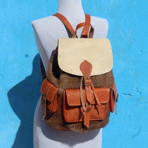 May include: A backpack featuring a combination of brown and cream-colored hair-on hide panels, accented with smooth tan leather pockets, straps, and a top flap. The bag includes a central buckle closure and two side pockets with metal hardware.