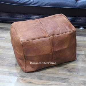 May include: A square, brown leather ottoman with visible stitching. The ottoman is placed on a wooden floor. The text "MoroccanBestShop" is visible on the side of the ottoman.