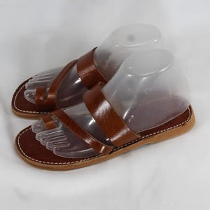 May include: A pair of brown leather sandals featuring a toe loop design and two horizontal straps across the top. The sandals have a flat sole with visible white stitching along the edge and are displayed on clear plastic foot forms.