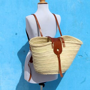 May include: A woven straw basket backpack featuring a wide, open-top design with a natural light beige finish. The bag includes brown leather shoulder straps and a central brown leather closure flap with a buckle-style strap detail.