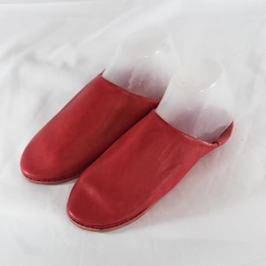 Puede incluir: Un par de pantuflas de cuero rojo con diseño de punta redondeada y estilo sin cordones. Las pantuflas se muestran con hormas de plástico transparente en el interior para mantener su forma, exhibiendo un acabado liso y costuras visibles.