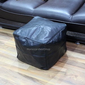 May include: A black leather pouf ottoman with a square shape. The pouf is sitting on a wooden floor.
