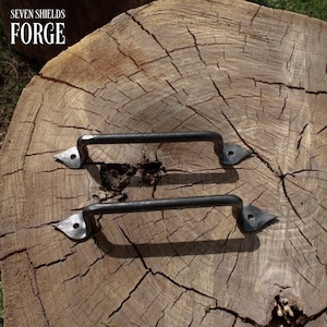 May include: Two black metal handles with decorative leaf-shaped ends are displayed on a weathered wooden surface. The handles have a curved design and are likely for a cabinet or drawer. The text "SEVEN SHIELDS FORGE" is in the upper left corner.
