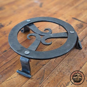 Op de afbeelding: Een handgemaakte, donkere metalen trivet met een cirkelvormig ontwerp en drie gebogen armen. De trivet heeft drie poten voor stabiliteit en is vastgeniet. Het Seven Shields Forge-logo is rechtsonder zichtbaar.