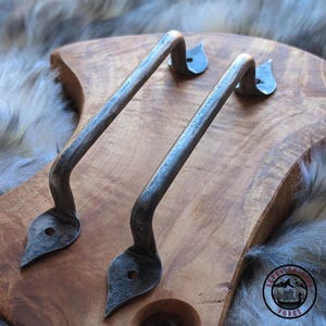 Puede incluir: Dos manijas de metal oscuro hechas a mano con un diseño curvo y extremos en forma de hoja. Las manijas están montadas sobre una superficie de madera, mostrando una estética rústica y forjada. Las manijas son probablemente para una puerta o cajón.