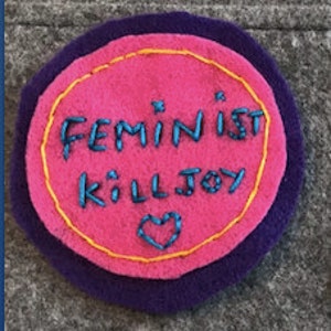 May include: A pink felt circle with a yellow border and purple backing. The circle has the text "Feminist Killjoy" embroidered in blue thread. A small heart is embroidered below the text.