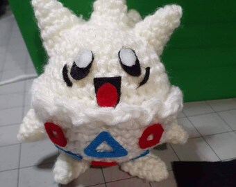 May include: White crocheted plush toy with red, blue, and black details. The toy has a smiling face with large eyes and a wide mouth. The toy is shaped like a round creature with a spiky head.