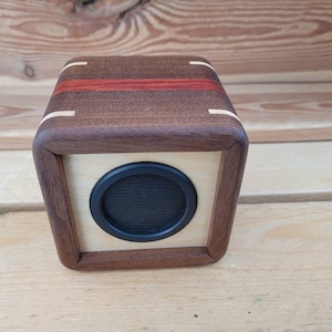 Op de afbeelding: Een kleine houten luidspreker met een zwart luidsprekerrooster. De luidspreker is gemaakt van donker hout met een lichte houten afwerking. De luidspreker staat op een houten oppervlak.