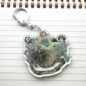 May include: A clear acrylic keychain featuring a cartoon illustration of four people standing on a stack of papers. The people are wearing different colored clothing and have different hairstyles. The keychain is attached to a white metal ring.