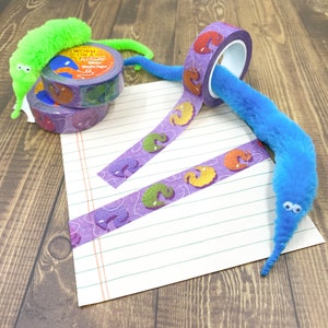 May include: Two rolls of purple and white patterned washi tape with the text "Worm on a String" and "Washi Tape" on one roll. The tape is unrolled and laid out on a sheet of lined paper. A blue fuzzy worm toy is wrapped around the tape and a green fuzzy worm toy is lying on the paper.